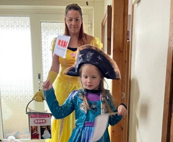 Abigail stands in front of her mum Deborah in the hallway of their home. Abigail is wearing a blue and purple princess dress with a black pirate hat. Abigail is waving a flag with the RSBC logo on it in each hand. Abigail's mum Deborah is standing behind her wearing a yellow princess dress and is holding an RSBC collection bucket in her right hand.