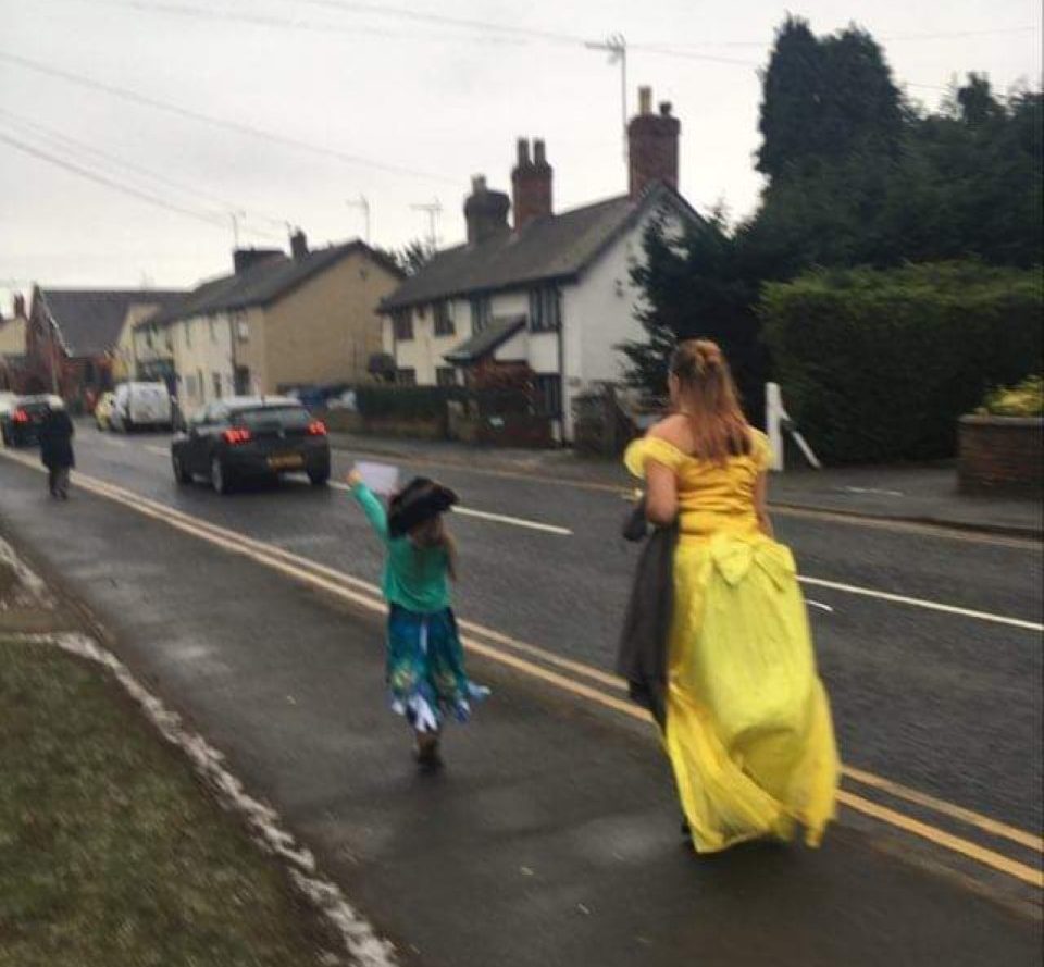 Abigail is running along the pavement a few steps in front of her mum, Deborah. Abigail is dressed in a blue and purple princess dress, a black pirate hat and a turquoise jumper. Deborah is wearing a yellow princess dress and carrying a grey jumper under her left arm. Abigail is waving her RSBC flag in her left hand at the cars that pass by.