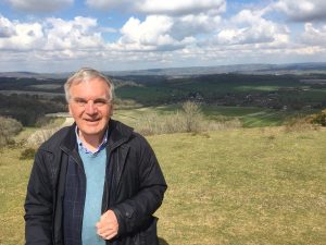 John is stood with a field behind him. The blue sky is dotted with clouds but he is wearing a coat.