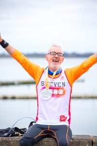 Steven is photographed holding his arms aloft and smiling, in front of the lake he ran around. He is wearing an RSBC running vest and has a homemade medal around his neck.