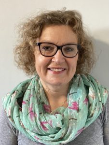 Sue Sharp standing against a white wall smiling, wearing glasses and a light blue flowery scarf