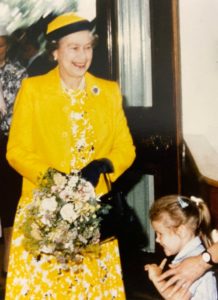 Her Majesty Queen Elizabeth smiling and holding flowers with a girl next to her