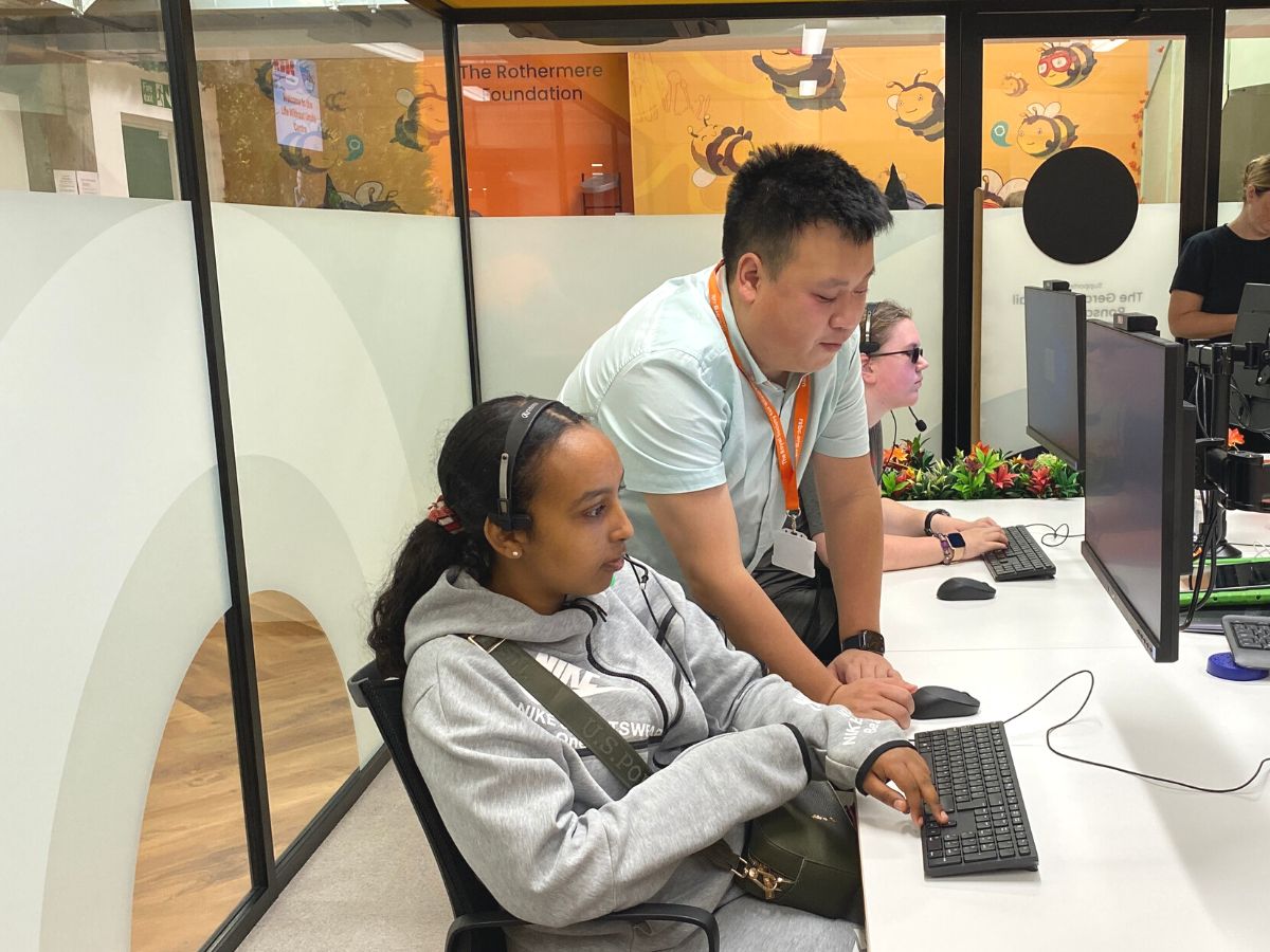 A woman sitting in front of a computer with a headset next to a man standing up and looking at the keyboard