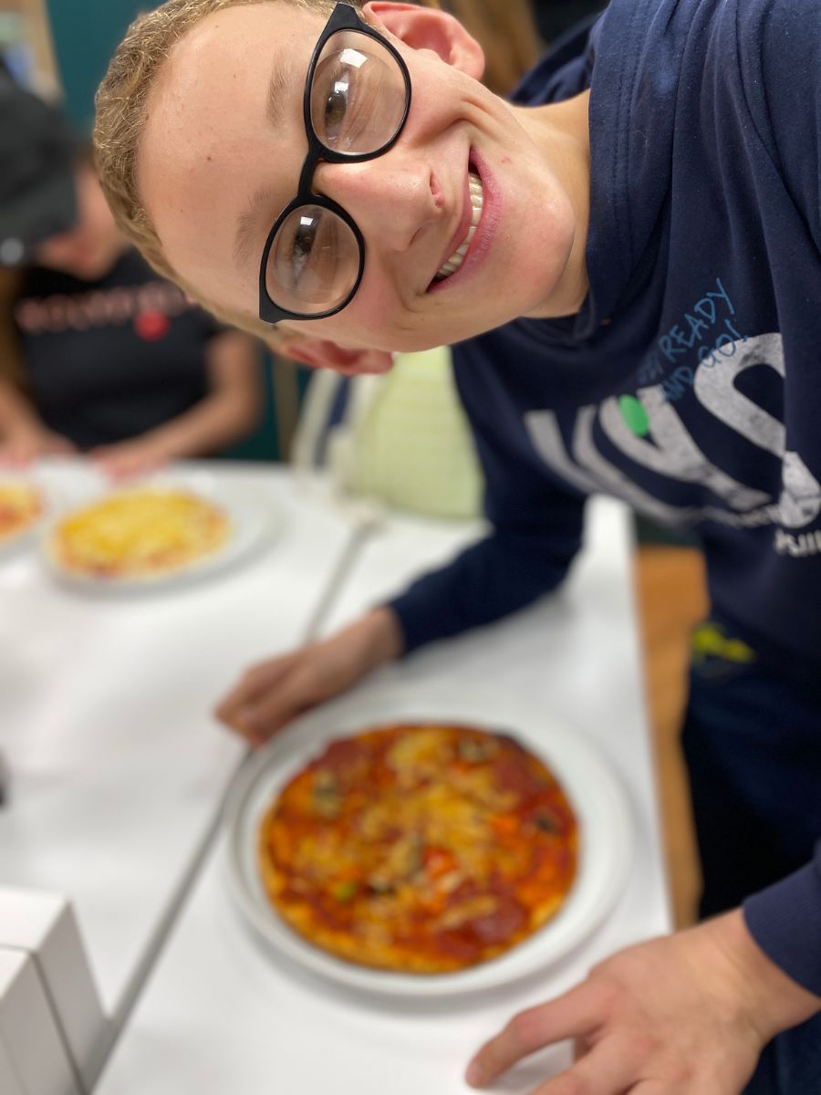 A young person in glasses smiling at the camera with a finished pizza on a plate