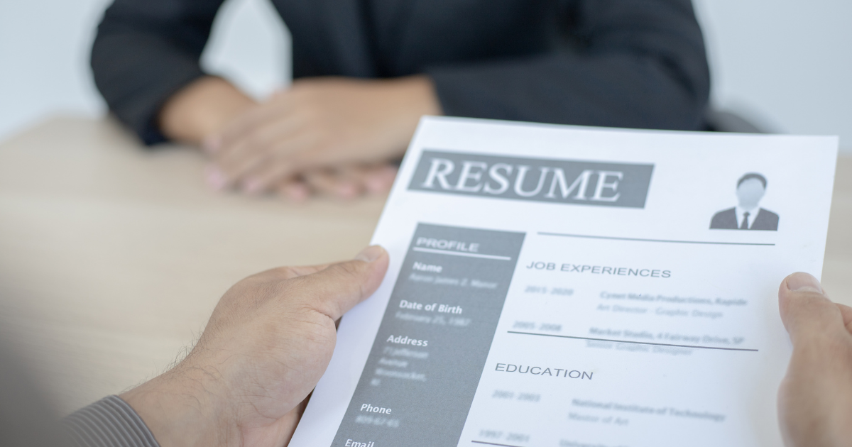 Two hands holding a document entitled 'Resume' with someone across the desk crossing their hands