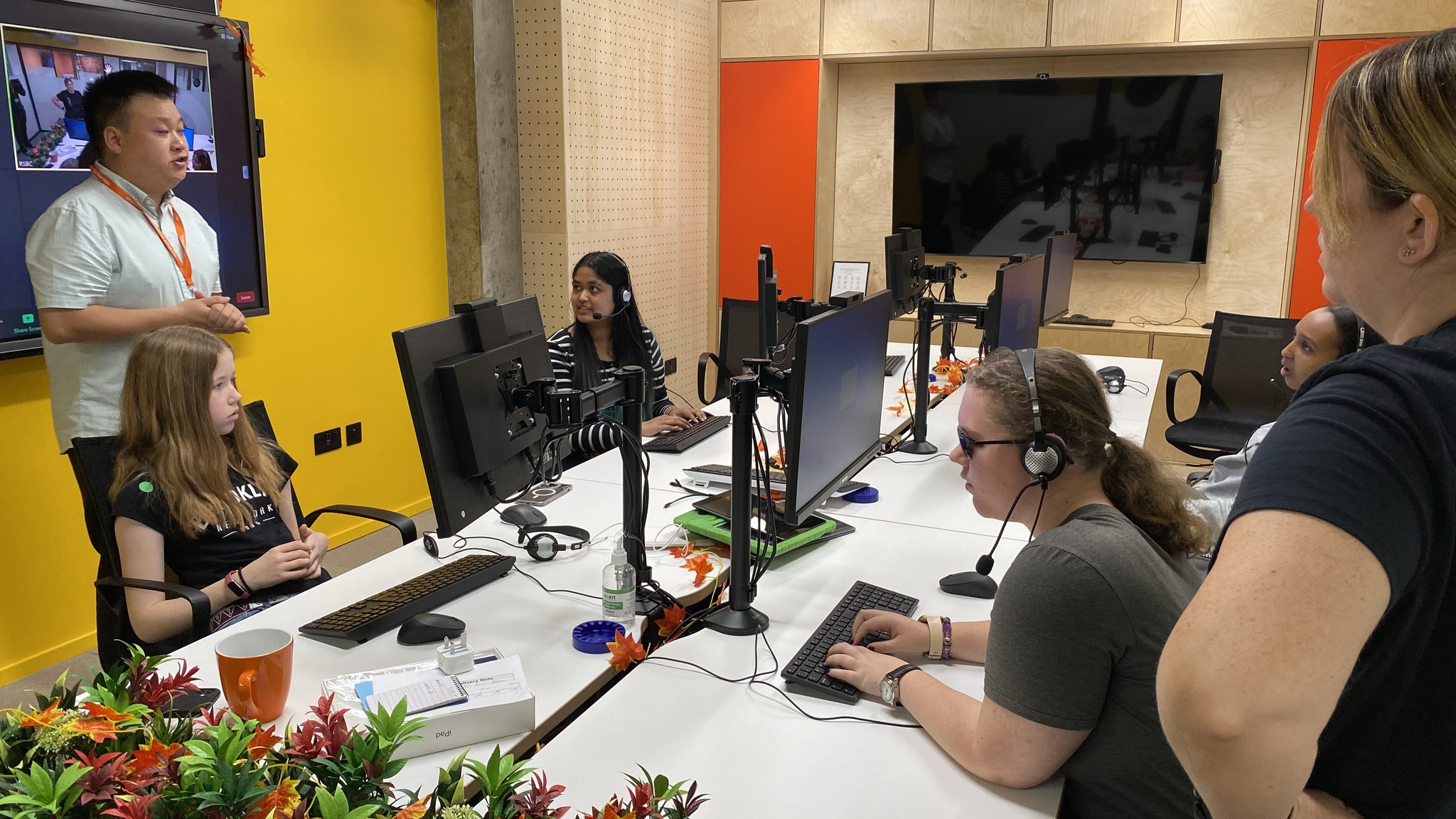 Young People in a room with computers listening to a man standing in front of a screen