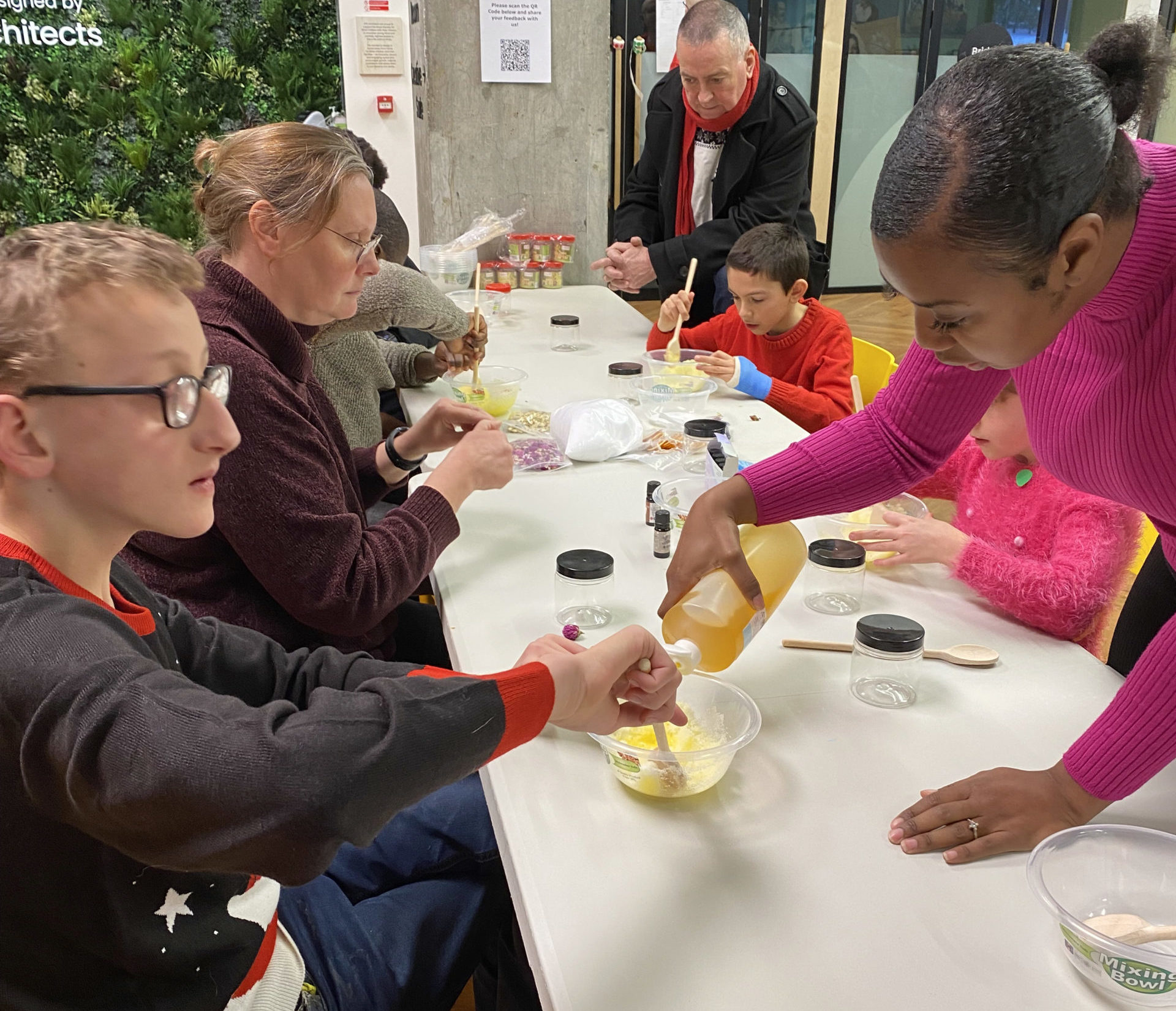 A group of people seating at a table with one woman pouring a liquid into a bowl and with a young boy mixing it.