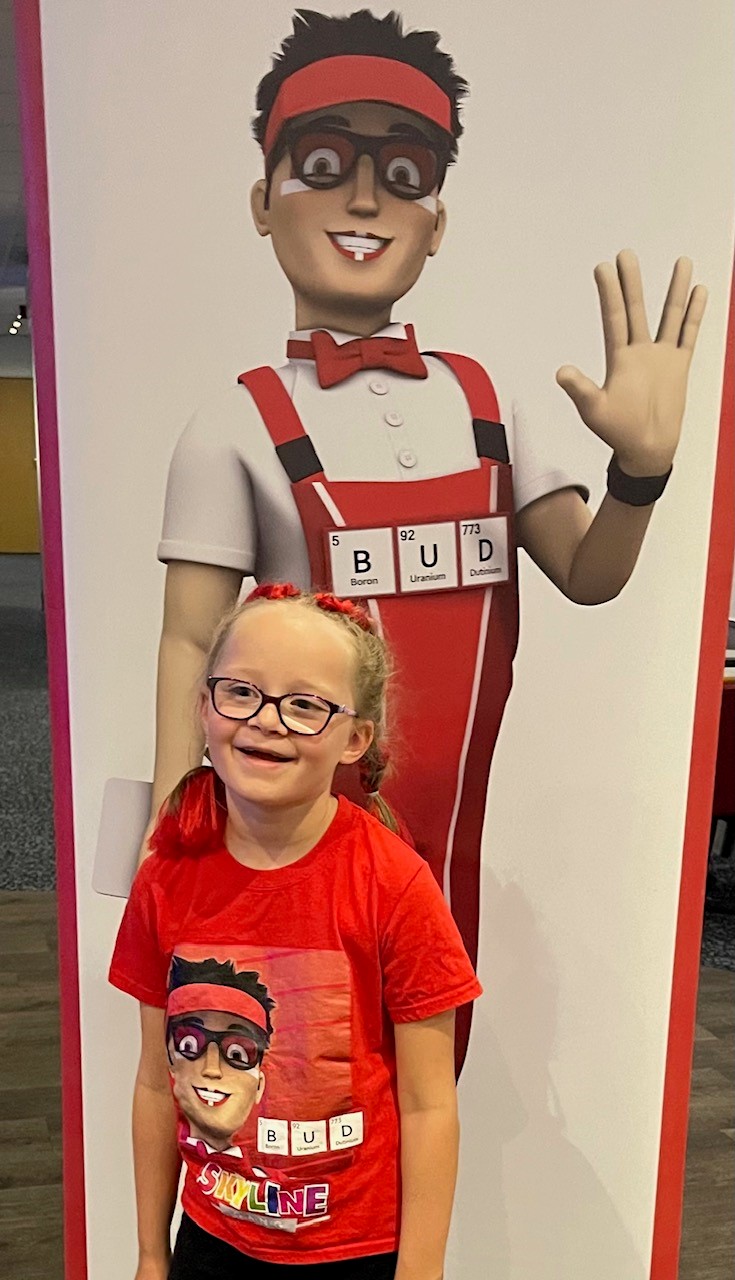 A young girl wearing glasses in a red outfit in front of a big poster.