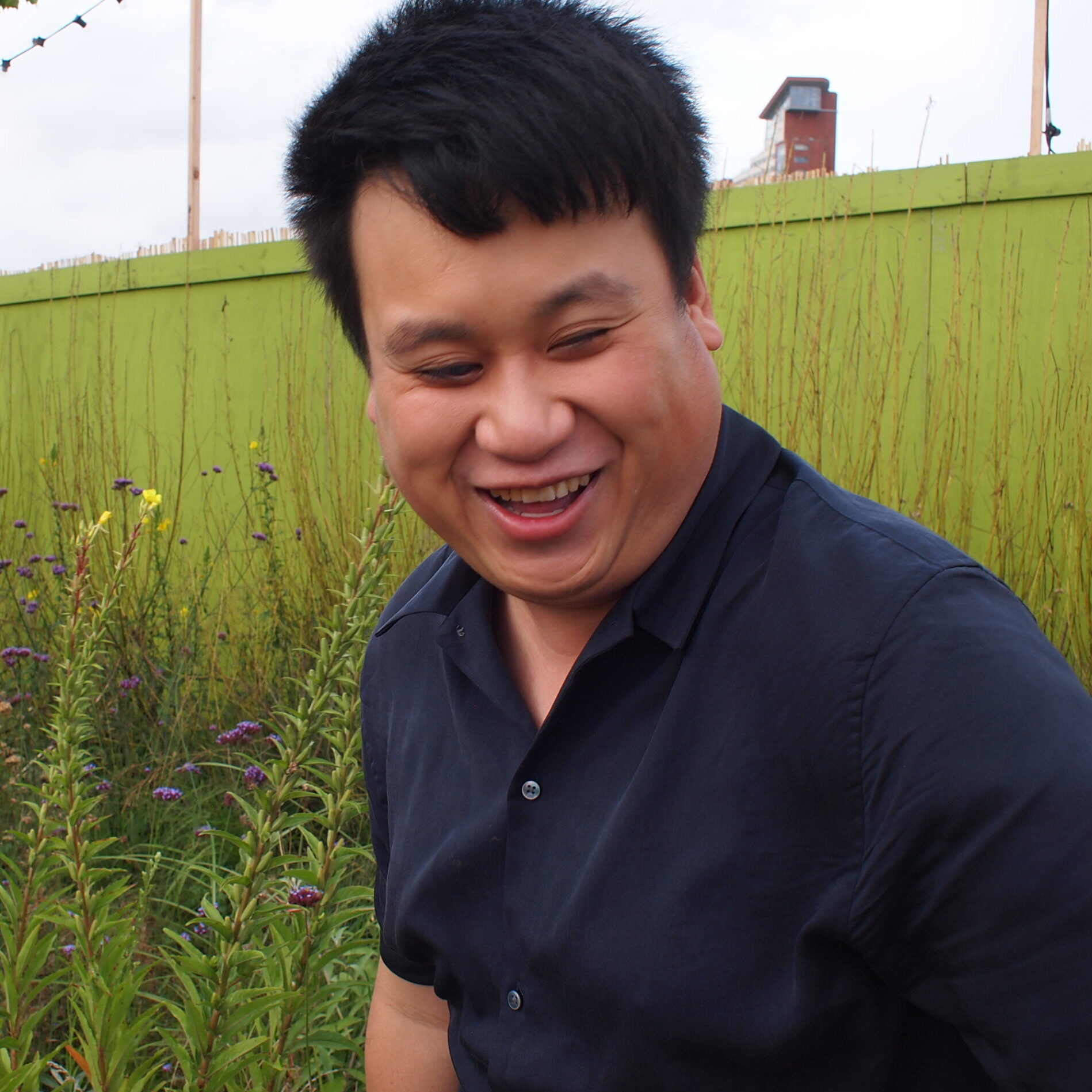 Alex, a man with thick black hair and a medium light skin tone, wears a blue shirt, laughing and looking downwards as he stands in front of a light green fence and foliage.