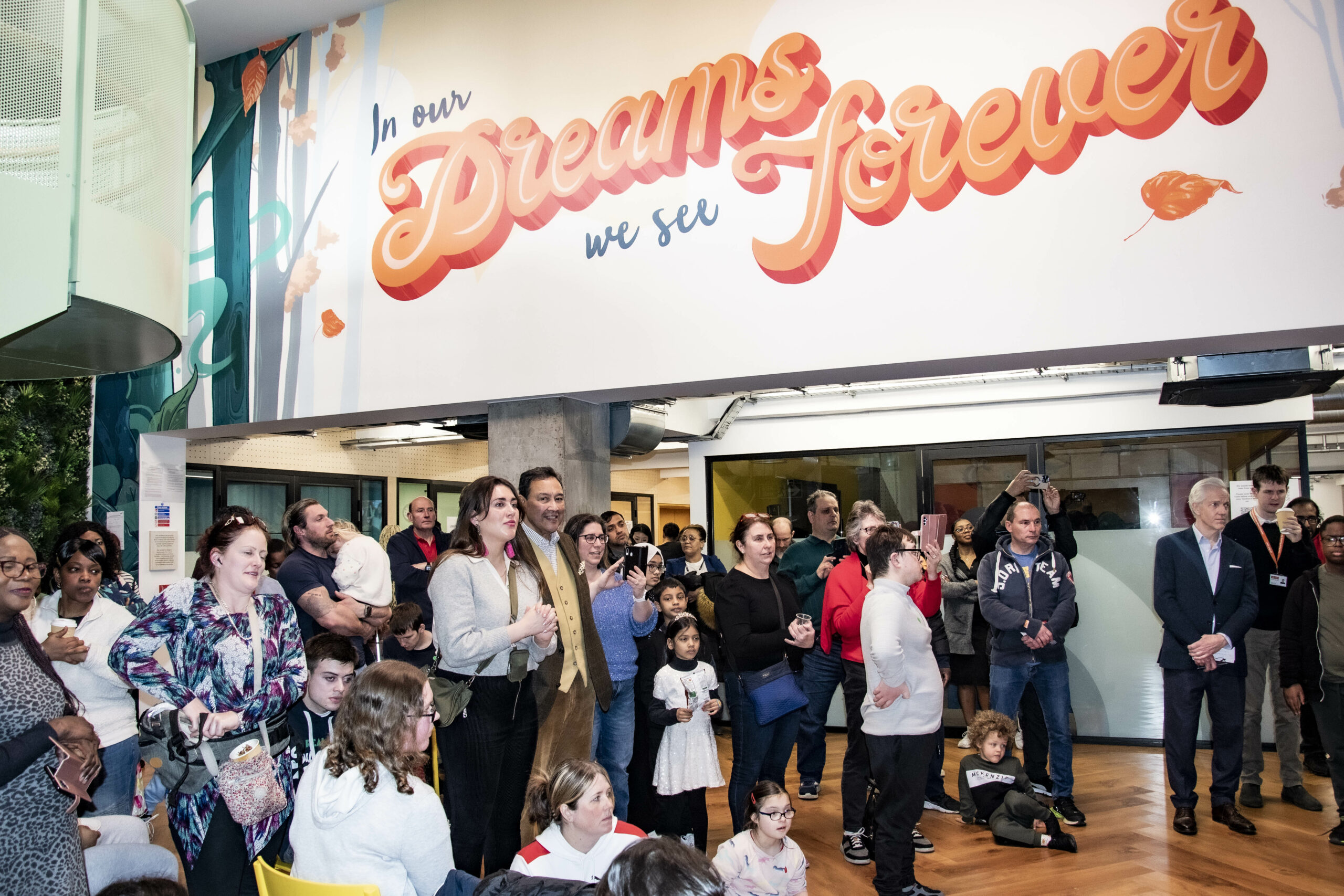 A group of people gathered in a big room underneath a sign that writes : "In our dreams we see forever"