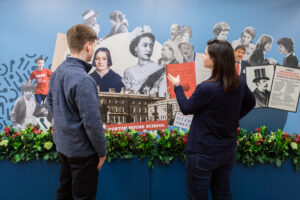 2 people standing up and looking at photos and illustrations of the Queen Elizabeth II on a wall