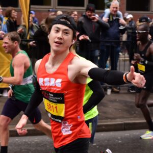 An RSBC London Marathon runner blazing past mile 23 with thumbs up. He is light-skinned with black hair, wearing an orange RSBC running vest.