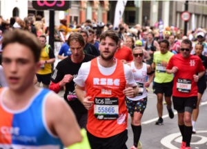 A street full of people running the London Marathon. The focus is on a man in the centre wearing a bright orange RSBC running vest.