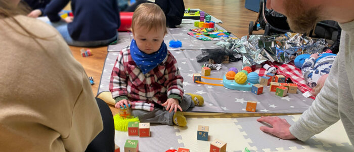 A baby sat on the floor surrounded by toys with parent sat in front of him looking at him