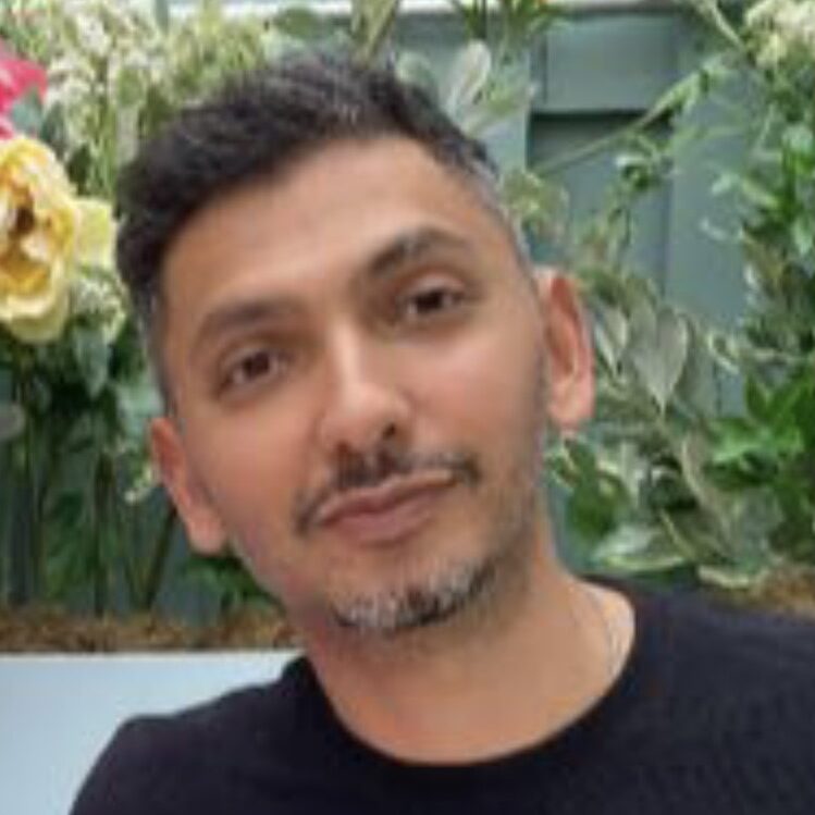 Vivek, a medium-skinned man with a short grey beard and hair wears a black T shirt and looks into the camera with a slight smile.