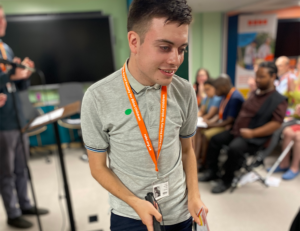 A young man with a white cane, holding his certificate. In the background is a room full of people, seated at the graduation ceremony.