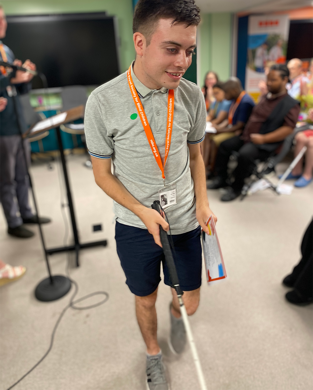 A young man with a white cane, holding his certificate. In the background is a room full of people, seated at the graduation ceremony.