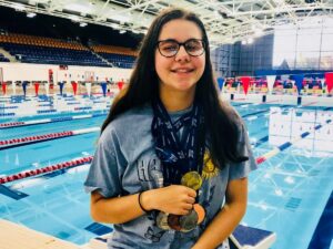 A young woman wearing glasses and several medals around her neck with a swimming pool in background.