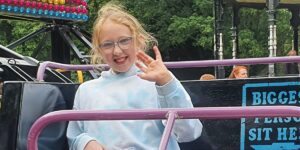 A light-skinned young girl wearing glasses at a fun fair and waving at the camera. She has blonde hair tied up in a ponytail and is wearing a light-blue hoodie.