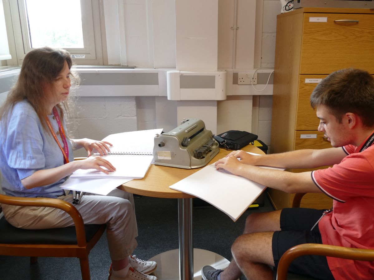 A woman reading braille at a table with a young man sitting in front of her . There is a braille machine on the table.