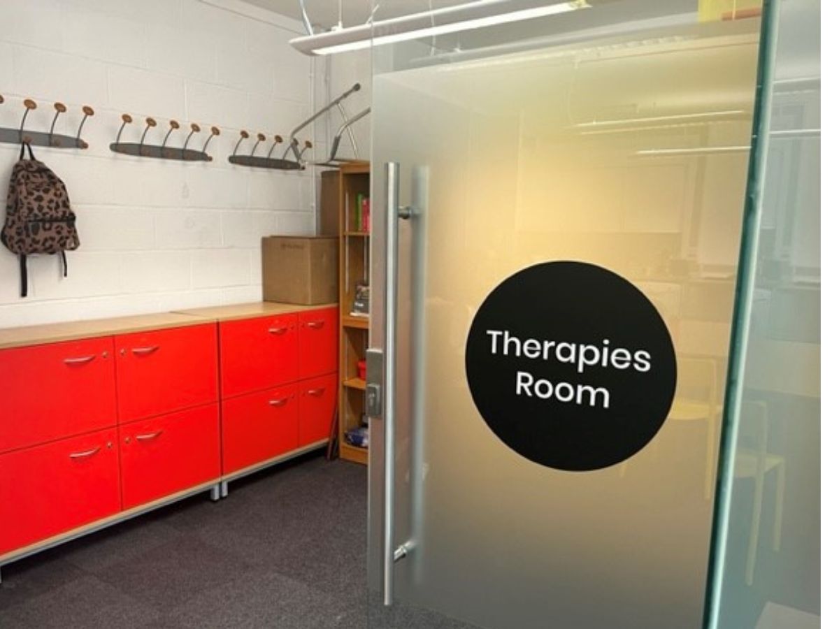 A room with a red cabinet and coat hangers. There is an open door with a sign that reads "Therapies room".