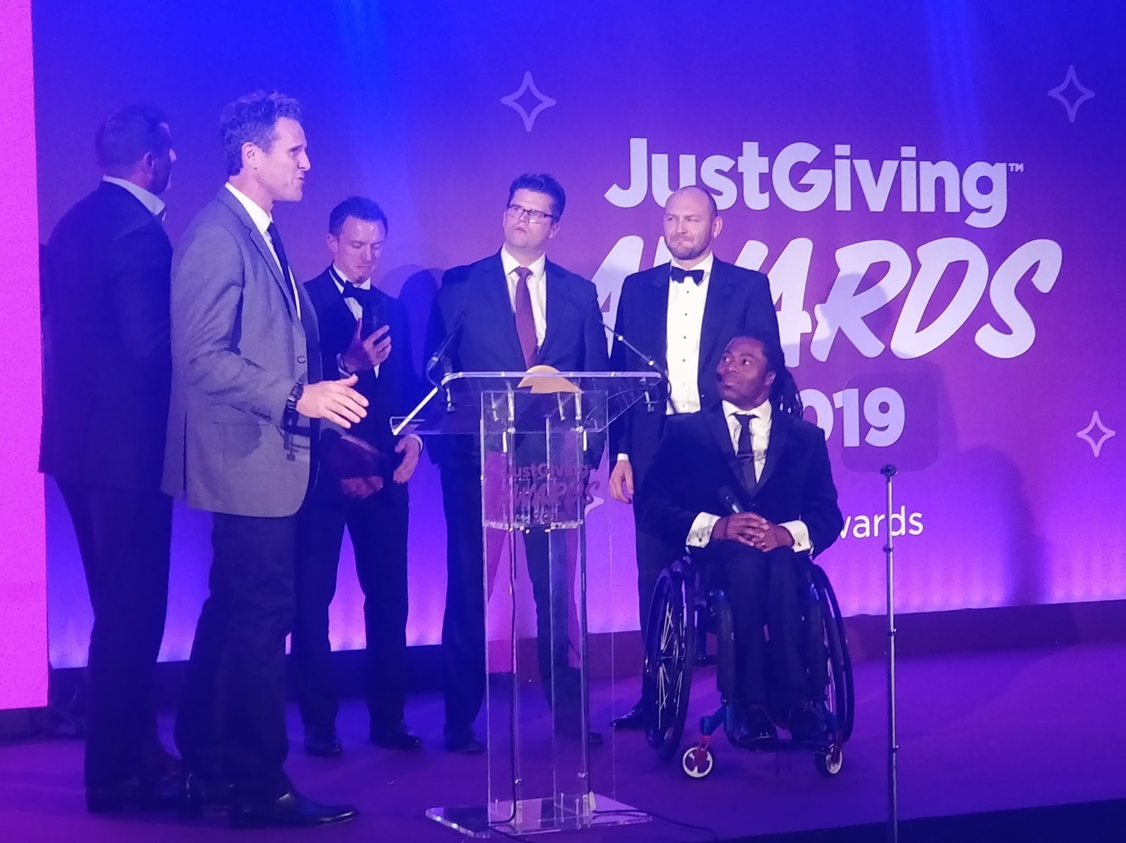 Six men dressed in formal wear on the stage at an awards ceremony. One of the 6 is TV Presenter - Ade Adepitan. In the photo far right presenting the award - James Cracknell, Olympic Rower