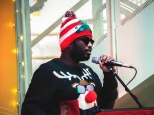 A young man wearing a Christmas jumper and hat standing up in front of a micro.