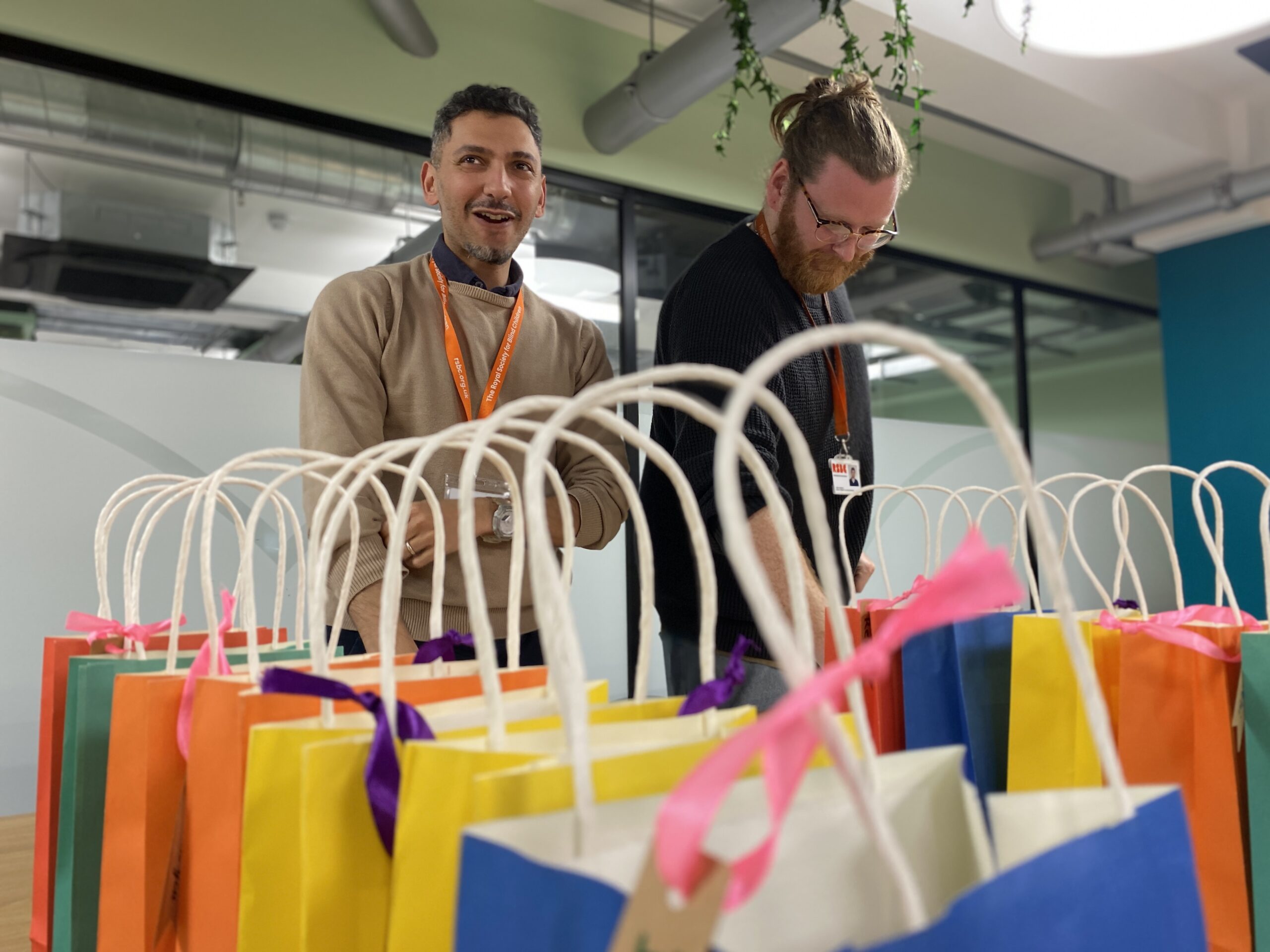 A number of colourful gift bags in the foreground, two people with lanyards in the background.