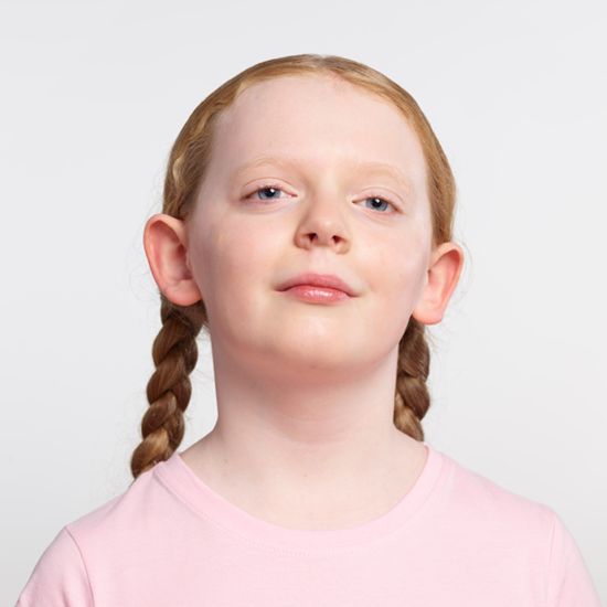 A blind girl stands against a white backdrop. She has red hair which is plaited and is wearing a pink t-shirt