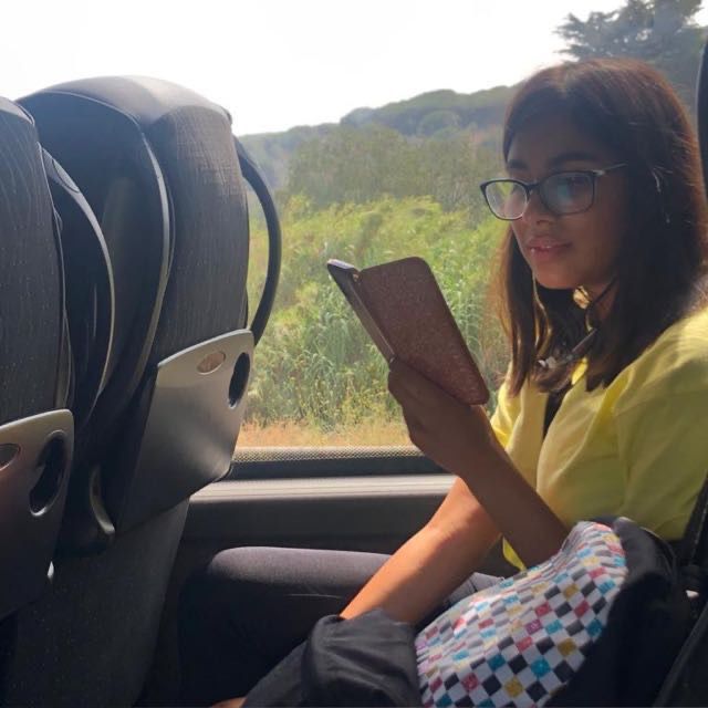 Alyssia, a medium light skinned person, sits on a train reading something intently on her mobile phone. She wears glasses and a yellow shirt and the countryside is visible out of the window beside her.