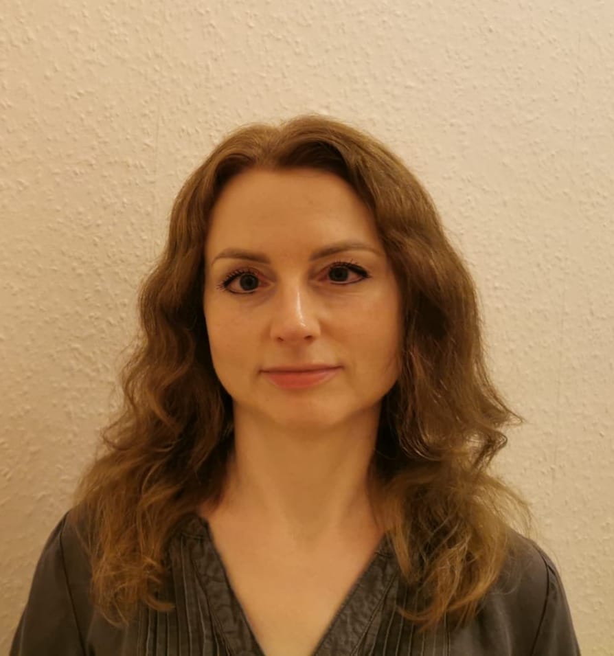 A head and shoulders shot of Edyta, a light skinned woman with long wavy mid-brown hair and a black top, looking straight ahead with a hint of a smile.