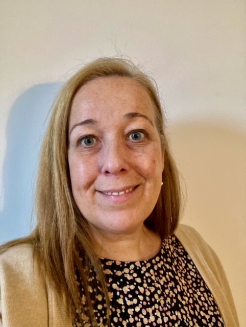 Jody, a woman with long straight blonde hair and a light skin tone, is wearing a patterned top with a fawn-coloured cardigan. She's smiling as she looks into the camera.