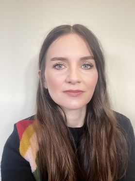 Justyna, a woman with long dark hair, light skin tone and light coloured eyes, wears a black top and looks at the camera with a serious expression.