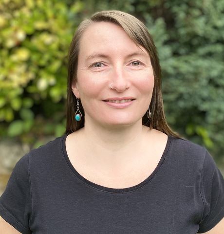 Nicola, a woman with light skin and long, straight brown hair, wears a black T shirt and turquoise earrings, and is looking straight ahead and smiling.