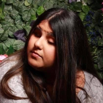 Zenny, a young person with long black hair and a medium skin tone, wears a grey and white patterned top. She looks serious as she looks away from the camera to the left.