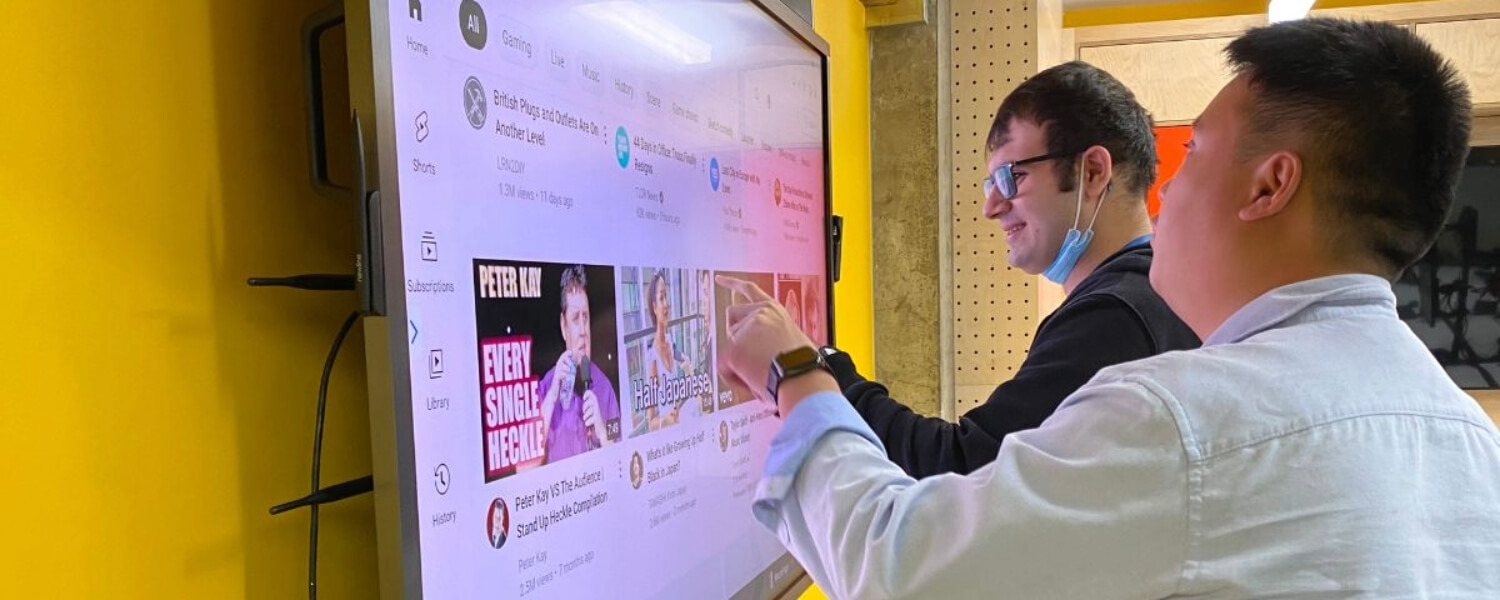 Two men standing in front of a big digital screen, close to the screen. The person farthest away from the camera has shirt brown hair and glasses. He is touching the screen with his finger. The person next to him is RSBC staff Alex. He has short brown hair and is guiding the other person's finger with his hand.