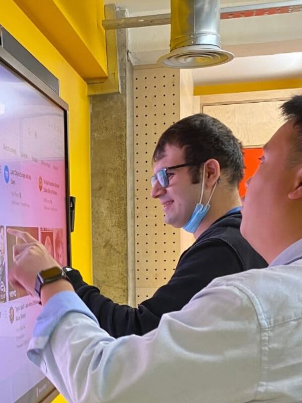 Two men standing in front of a big digital screen, close to the screen. The person farthest away from the camera has shirt brown hair and glasses. He is touching the screen with his finger. The person next to him is RSBC staff Alex. He has short brown hair and is guiding the other person's finger with his hand.