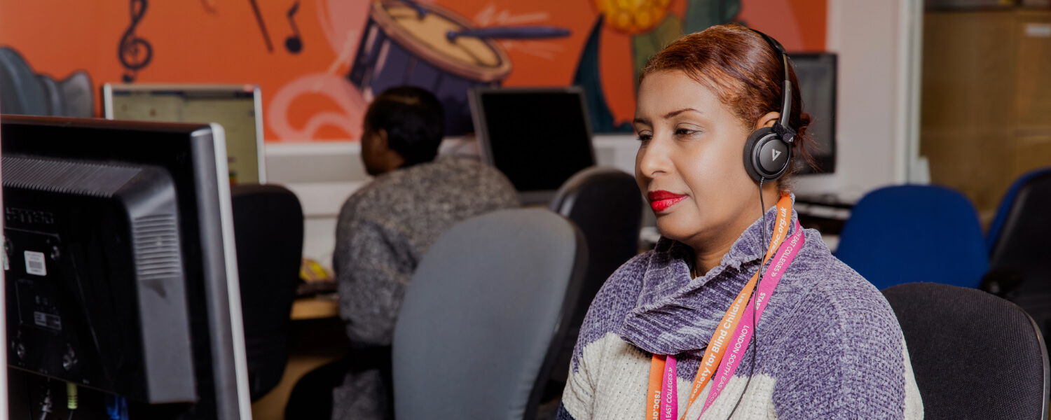 A medium skinned RSBC employee with red hair wears headphones and sits at a desk looking at a computer screen and keyboard. Other employees are in the background.