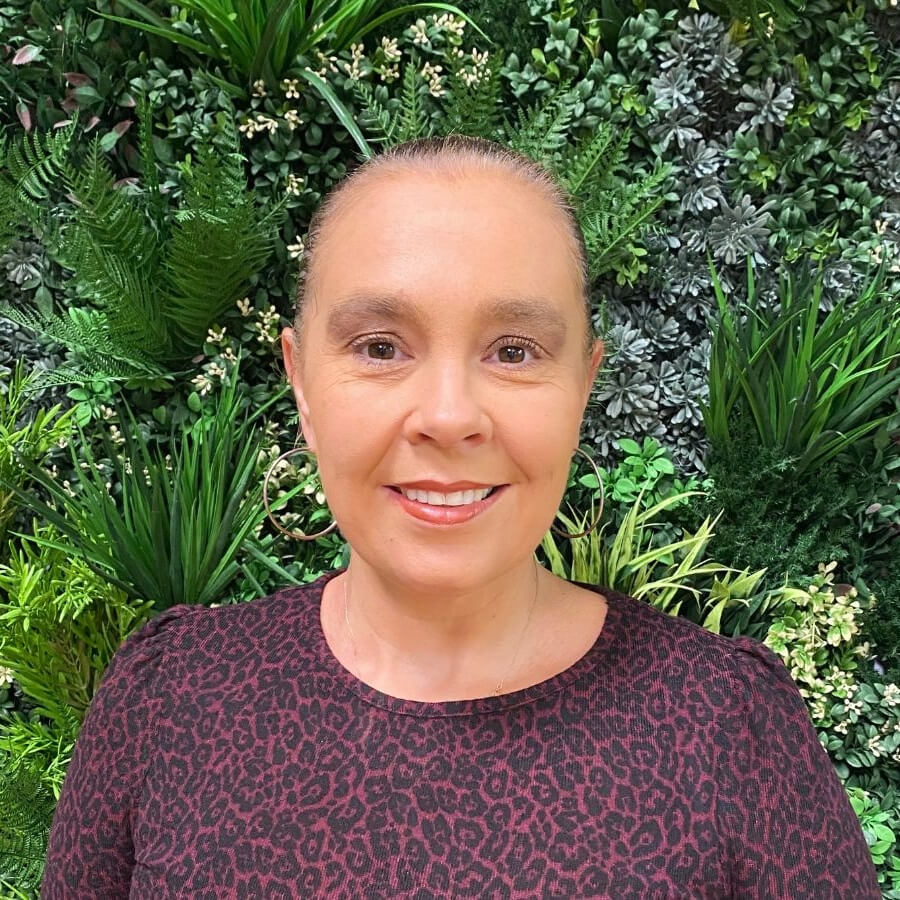 Carla, light skinned woman is smiling warmly at the camera. Her hair is tied tightly back and she wears a dark pink and black patterned top as she stands in front of a wall of green foliage.