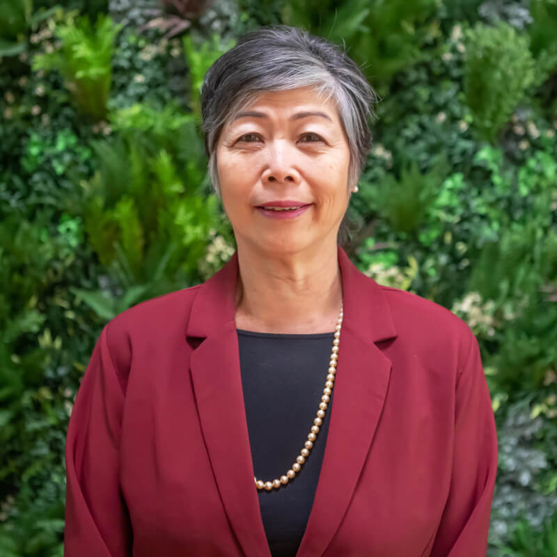 Eileen, a medium-light skinned woman with short grey hair stands in front of a wall of lush green foliage. She's smiling, and wears a burgundy coloured jacket, black top and long gold necklace.