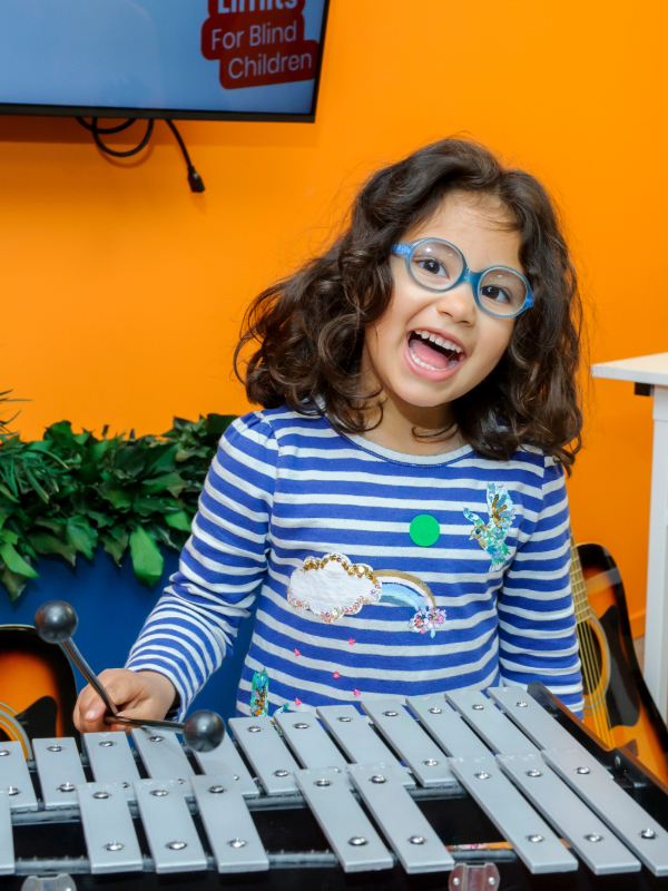 A young dark haired girl with medium light skin tone wears blue glasses and smiles at the camera as she plays the xylophone. The wall behind her is orange, and there's some green foliage behind her too.