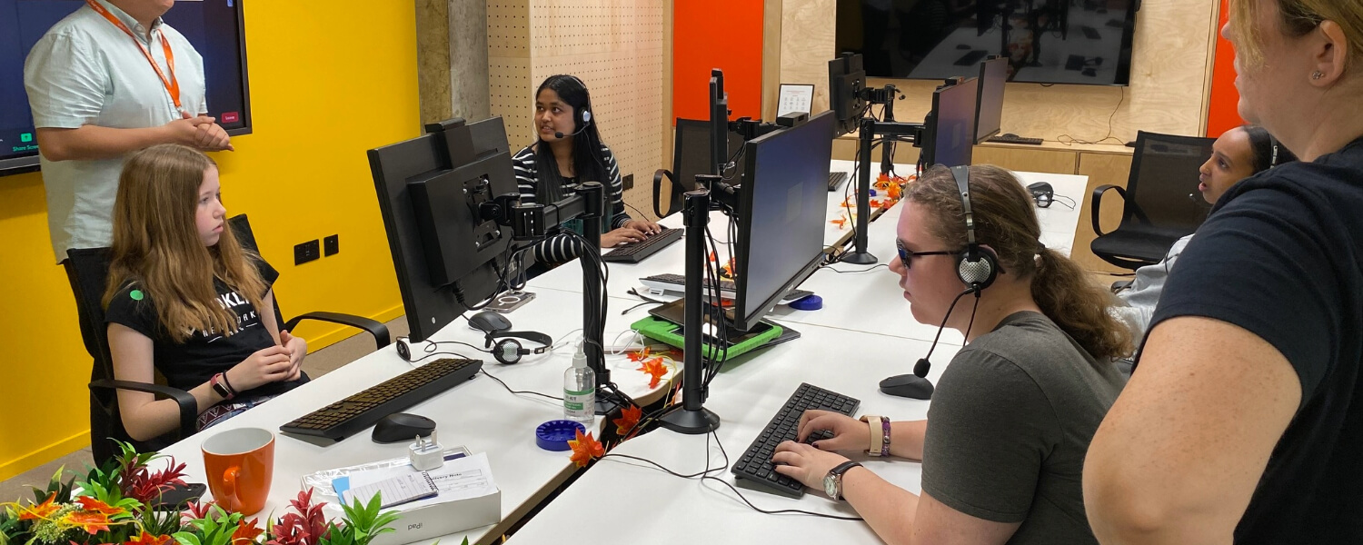 Young People in a room with computers listening to a man standing in front of a screen