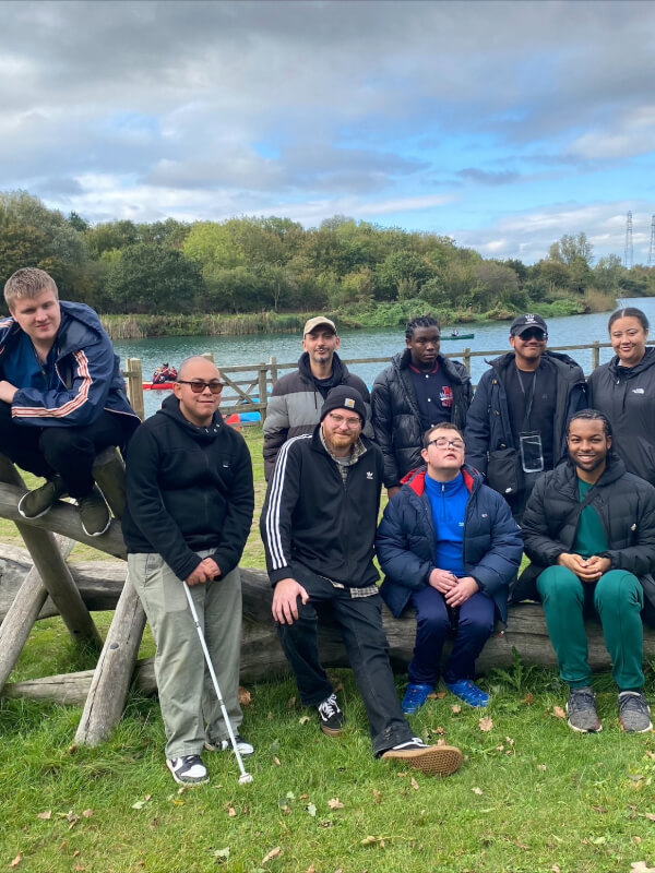 A group of young people with different skin tones stand and sit outside, in front of a wooden climbing frame with an expanse of water in the background. They are smiling at the camera and one in the front row holds a white cane.