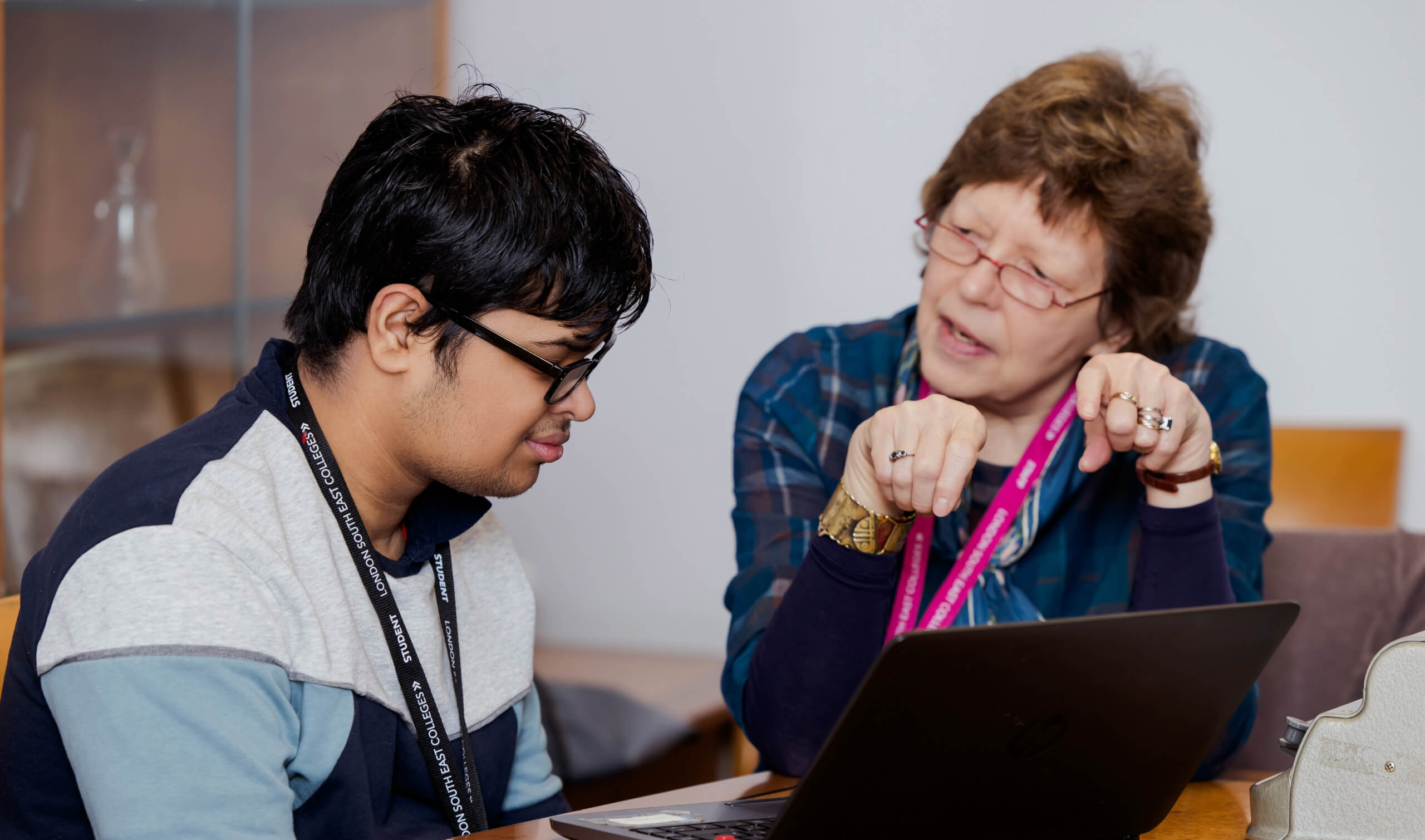 An RSBC staff member with a light skin tone is wearing glasses, a blue top and a pink lanyard. She sits at a table talking to a dark haired, medium skinned student in a blue and white top. They sit in front of a laptop.