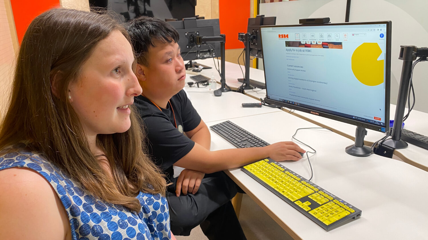 A young man and woman sit in front of a PC screen, using a yellow keyboard