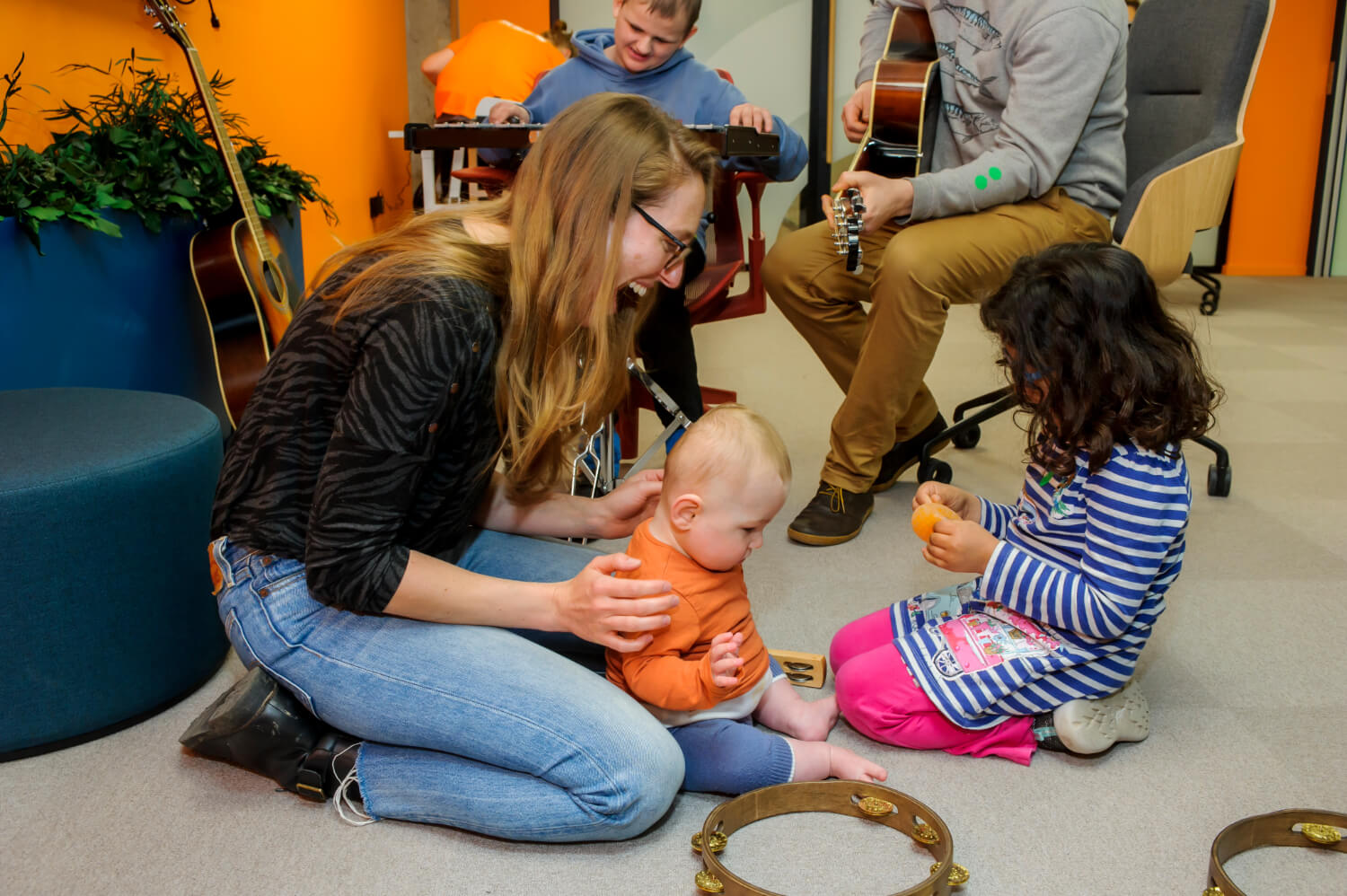 A mum sits on the floor holding her small child while a young girl plays in front of them