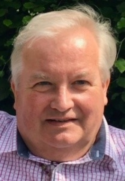 Ian, a light skinned man with light grey hair wears a shirt with small white and red checks. He is looking directly at the camera and a slight smile is on his lips.