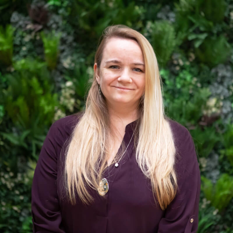 Josie, a light skinned woman with long blonde hair, is smiling at the camera. She wears a purple top and is standing against a wall of green foliage.