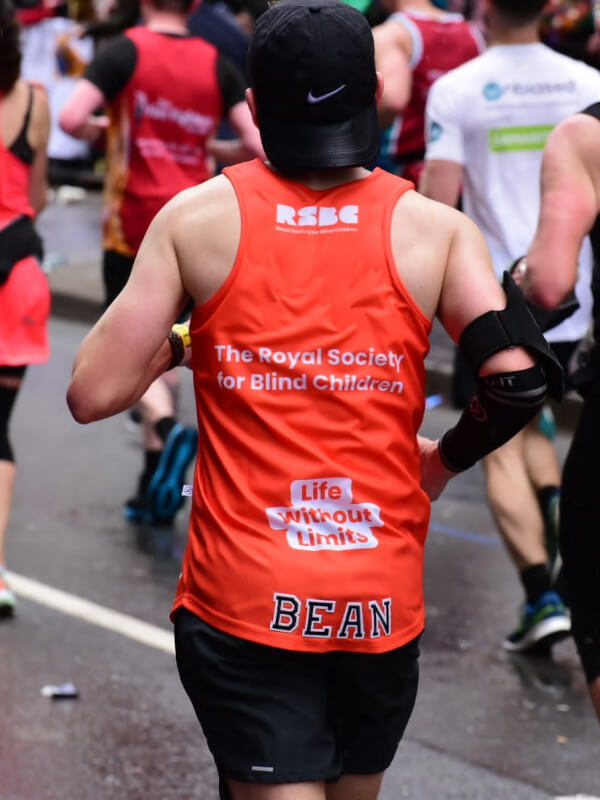 The back of a light skinned runner at the London Marathon. He is wearing a branded RSBC orange running top which says 'BEAN' at the bottom.