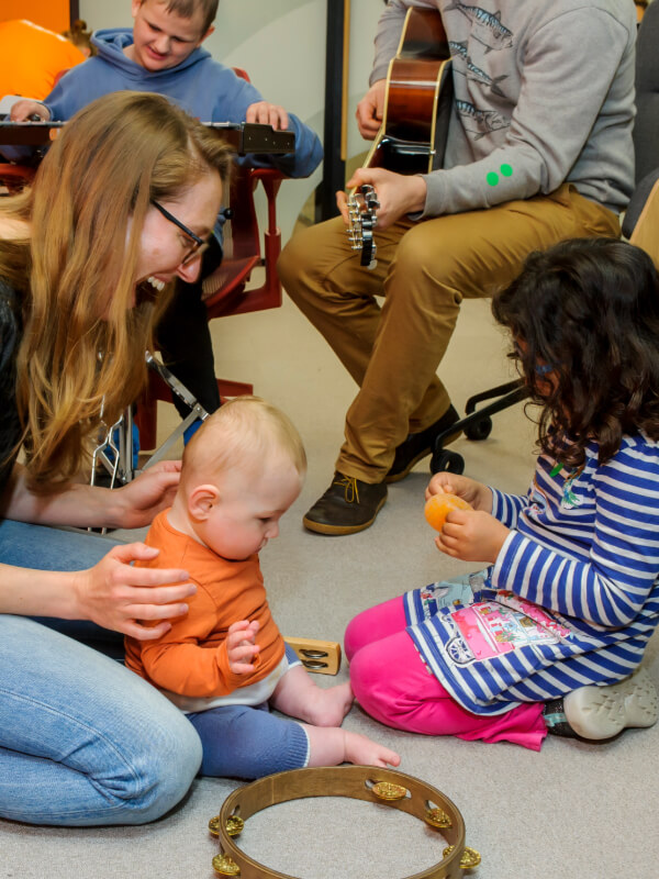 A mum with long blonde hair kneels on the floor holding her little boy who has an orange top on. A young girl with long brow hair and a clue and white stripey dress and bright pink trousers plays beside them